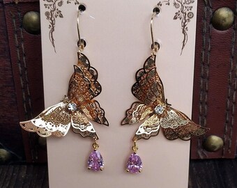 Puede incluir: Un par de pendientes de mariposa dorados con intrincados detalles calados. Cada pendiente presenta una gema transparente y una gema púrpura en forma de lágrima. Se exhiben en una tarjeta rosa con el sitio web www.steamspirits.net.