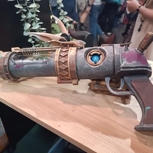 May include: A steampunk-style prop gun with a gray and brown color scheme. The gun features intricate details, including a glass tube, decorative bands, and a blue orb. The handle is a dark brown color.