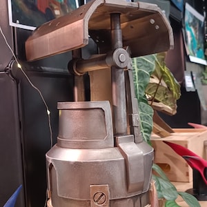 Puede incluir: Escultura metálica de color bronce con base cilíndrica y parte superior curvada. La escultura está montada sobre una base de madera. En primer plano, una escultura abstracta separada en tonos marrón y dorado.