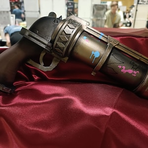 May include: A prop gun with a dark brown handle and a metallic barrel. The barrel is painted gold and has blue and pink paint splatters. The gun is resting on a red satin surface.