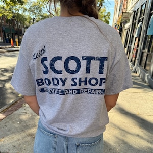 Pode incluir: Uma camiseta cinza com o texto azul marinho "Keith SCOTT BODY SHOP SERVICE AND REPAIR". A camiseta é usada com jeans azul claro. A imagem é tirada ao ar livre.