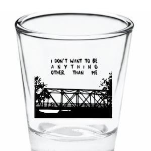 May include: A clear glass shot glass with a black silhouette of a bridge and the text "I don't want to be anything other than me".