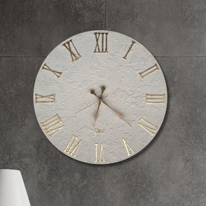 May include: A white round clock with Roman numeral markings and gold-colored hands against a gray textured wall. The clock is hanging above a wooden table with a lamp and books.