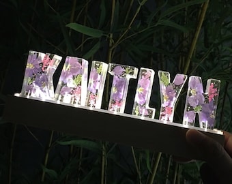 May include: A lighted sign with the name "Kaitlyn" spelled out in clear acrylic letters. Each letter is filled with pressed purple flowers.