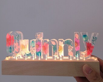 May include: A wooden base with a light-up name sign that says "Quinn" in clear resin with embedded pressed flowers. The flowers are pink, blue, and yellow.