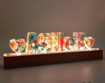 May include: A wooden base with a light-up display featuring the name "Ashley" spelled out in clear resin letters filled with colourful pressed flowers. The letters are illuminated from below, creating a warm glow.