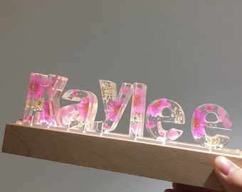 May include: A custom name sign with the name "Kaylee" spelled out in clear resin letters. Each letter is filled with pink pressed flowers and gold glitter. The letters are mounted on a wooden base.