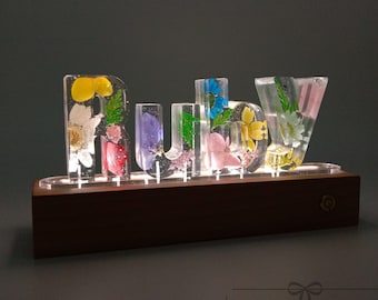 May include: A clear glass name sign with the word "Ruby" spelled out in large letters. Each letter is filled with pressed flowers in various colours, including pink, yellow, blue, and green. The sign is lit from below and sits on a wooden base.