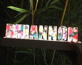 May include: A white wooden sign with the word "Rhiannon" spelled out in clear plastic letters. Each letter is filled with colorful pressed flowers.