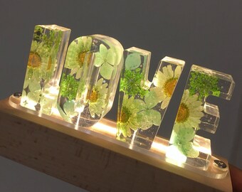 May include: A wooden base with a light-up sign spelling "LOVE" in clear resin with pressed green flowers and leaves.