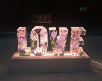 May include: A white wooden base with a light-up sign spelling "LOVE" in clear acrylic letters. Each letter is filled with pressed flowers in shades of pink and purple.