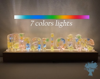 May include: A personalised name sign with the name "Charlotte" spelled out in clear resin letters. Each letter is filled with pressed flowers and illuminated with 7 colour lights. The sign is on a wooden base.