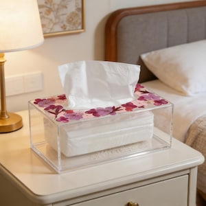 May include: A clear acrylic tissue box with a floral design. The rectangular box holds a stack of white tissues. The top is decorated with pink and purple flowers. The box sits on a white bedside table.