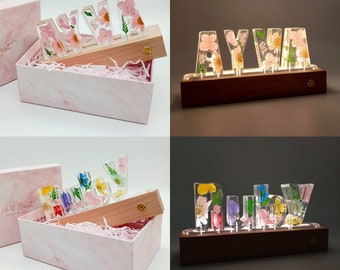 May include: Four different custom-made light-up name signs with pressed flowers in clear resin. The names are "XYLA", "AYVA", "Indy", and "Ruby". Each name is on a wooden base with a light switch on the side.