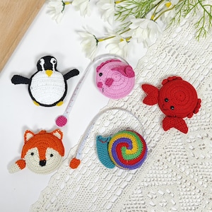 May include: A selection of crocheted animal measuring tapes. The set features a penguin, fox, pig, fish, and snail. The tapes are white with black, orange, pink, red, and rainbow details. Each tape has a pink or orange pull tab.