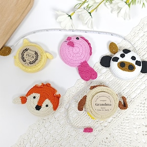 Puede incluir: Una colección de cintas métricas de ganchillo con forma de animales, incluyendo un pez amarillo, un pez rosa, una vaca, un zorro y una cinta beige con el texto "Abuela". Cada cinta tiene un diseño y una combinación de colores diferentes, con una cinta métrica que se extiende desde cada una.