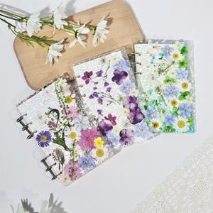 Op de afbeelding: Drie transparante notitieboekjes met zilveren ringbanden, versierd met geperste bloemen in paars, roze, geel en blauw. De notitieboekjes liggen op een wit oppervlak, met een houten plank en witte bloemen. Het bloemendesign is een belangrijk kenmerk.
