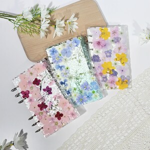Op de afbeelding: Drie transparante harsnotitieboekjes met geperste bloemen in roze, blauw en geel. Elk notitieboekje heeft een zilveren spiraalbinding. De notitieboekjes zijn gerangschikt op een wit oppervlak met een houten snijplank en bloemaccenten.
