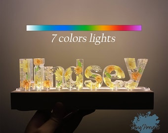 May include: A personalised name sign with the name "Lindsey" spelled out in clear acrylic letters. Each letter is filled with pressed flowers and illuminated with LED lights that change colour. The sign is on a wooden base and has a 7 colour light option.