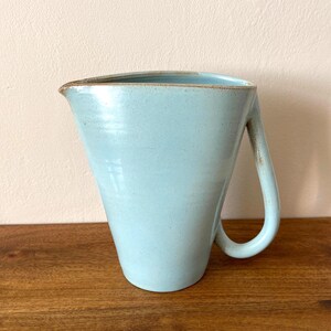 May include: A light blue ceramic jug with a wide, flared top and a single curved handle. The vessel features a smooth, matte finish with subtle speckling throughout and a distinct, slightly darkened rim along the top edge and handle.