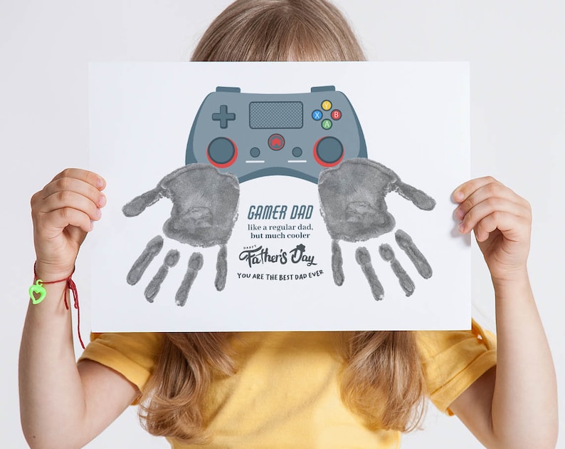 K&ouml;nnte beinhalten: Ein Kind h&auml;lt eine wei&szlig;e Karte mit einem grauen Handabdruck und einer Zeichnung eines Videospiel-Controllers hoch. Die Karte sagt "Gamer Dad wie ein normaler Vater, aber viel cooler. Frohes Vatertags. Du bist der beste Papa der Welt."