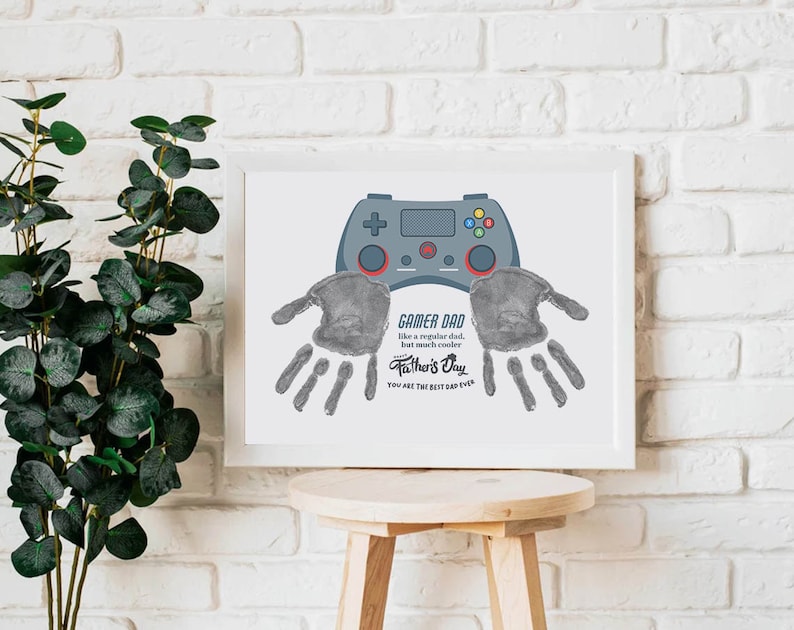 K&ouml;nnte beinhalten: Ein wei&szlig;er gerahmter Druck mit einem grauen und blauen Videospiel-Controller und zwei grauen Handabdr&uuml;cken. Der Text lautet "Gamer Dad like a regular dad, but much cooler. Happy Father's Day. You are the best dad ever."