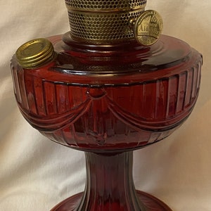 Może przedstawiać: Czerwona szklana lampa naftowa z mosiężnym palnikiem i złotą obwódką wokół podstawy. Lampa ma dekoracyjny wzór z drapowanej tkaniny wokół środka.
