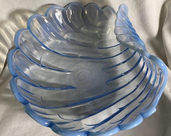 Puede incluir: Un cuenco de vidrio azul claro con forma de concha. El cuenco tiene un borde festoneado y una serie de líneas curvas que crean un patrón similar a una concha. El cuenco está hecho de vidrio transparente con un tinte azul claro.