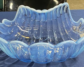 Puede incluir: Un cuenco grande de vidrio azul opalino con un diseño festoneado en forma de concha. El cuenco tiene una forma amplia y abierta con una serie de crestas curvas y un acabado liso y brillante. El color es un azul suave y translúcido.