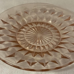 May include: A clear, pink glass plate with a diamond-cut pattern. The center features a sunburst design. The plate is round and has a slightly raised edge. This vintage dish is suitable for serving small portions.