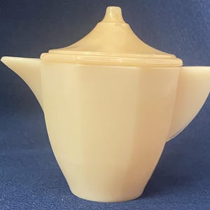 Op de afbeelding: Een crèmekleurige, ondoorzichtige glazen theepot met een gefacetteerd, geometrisch lichaam, een puntig deksel, een driehoekige tuit en een bijpassend handvat. Het object heeft een gladde, glanzende afwerking en een consistente lichtbeige kleur.