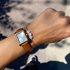 May include: A gold watch with a white face and brown leather band. The watch is on a person's wrist with a red string bracelet with white beads that spell "THV".