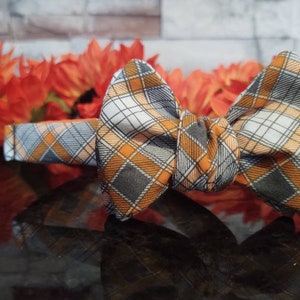 May include: A bow tie with a grey, white and orange plaid pattern. The bow tie is made of fabric and has a classic bow shape.