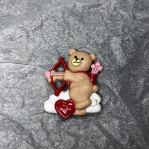 May include: A Valentine's Day themed figurine of a teddy bear dressed as Cupid, holding a red bow and arrow. The bear is tan with a red scarf and is standing on a white cloud. A red heart with the words "Be Mine" is at the bottom.