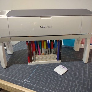 May include: A white Cricut Maker cutting machine with a gray top and a silver trim. The machine is on a gray cutting mat with a ruler and grid pattern. There are a variety of colored markers in a holder next to the machine.