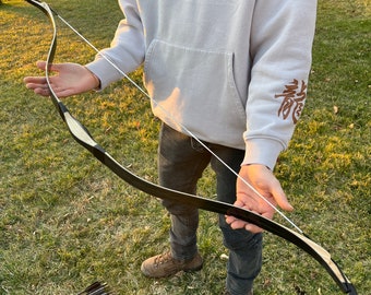 May include: A person holding a black and wooden bow with a white string. The bow is about 60 inches long. The person is wearing a grey hoodie with the word "SHOUT" on it and black jeans.