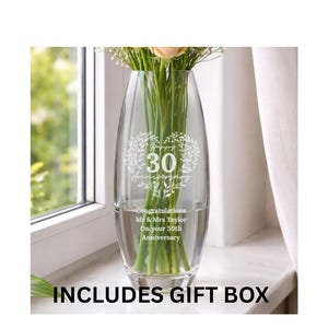 May include: A clear glass vase with a tapered oval shape, featuring an etched heart-shaped wreath design surrounding the number 30. Below the wreath, the text reads: Congratulations Mr & Mrs Taylor On your 30th Anniversary. Green flower stems are visible inside.