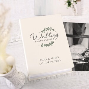 May include: A white wedding photo album with a green leafy design and the text "Wedding Photo Album" and "Emily & James 14th April 2023".