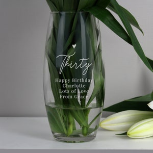 May include: A clear glass vase with a curved shape, containing water and green foliage. The vase has the word "Thirty" in cursive script, plus a birthday message. White lilies are seen in the background.