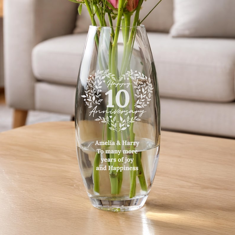 May include: A clear glass vase featuring an etched design of a leaf wreath surrounding the text Happy 10 Anniversary. Below the wreath, the text reads Amelia & Harry To many more years of joy and Happiness. The vase contains green flower stems.