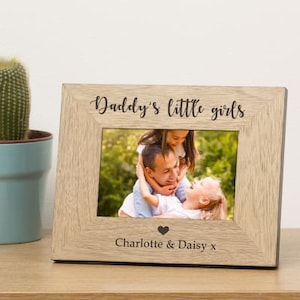 May include: A light wood photo frame with a black inscription that reads "Daddy's little girls" and a heart symbol. The frame has a photo of a father with his two daughters. The inscription below the photo reads "Charlotte & Daisy x".