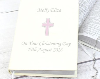 May include: Cream-colored book with the text "Molly Eliza" and "On Your Christening Day 19th August 2026." A pink cross is centered on the cover. A rosary and an open book are in the background.