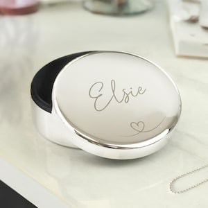 May include: A silver-coloured, round jewellery box with the name "Elsie" engraved on the lid, along with a small heart. The box is open, revealing a black interior. The box is on a white surface.
