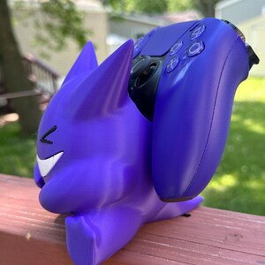 Peut inclure: Un support de manette de jeu violet en forme de personnage de dessin anim&eacute;, tenant une manette violette assortie. Le support a un visage souriant et des oreilles pointues. La manette est de couleur violet fonc&eacute;.