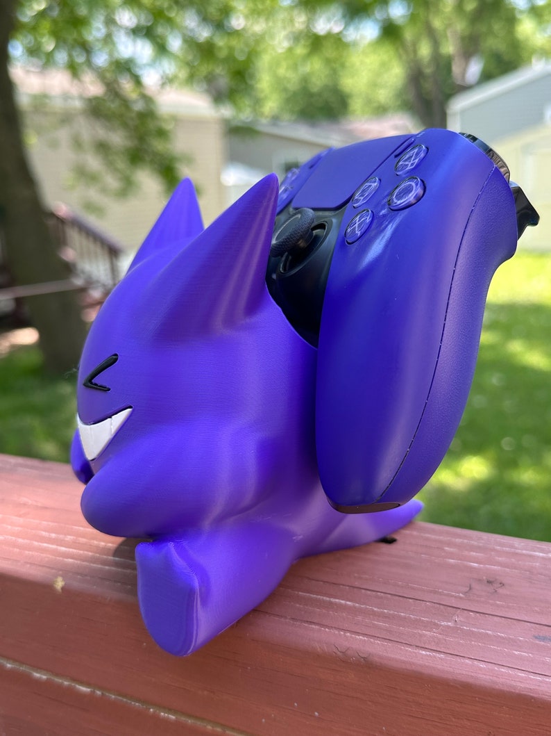 Peut inclure: Un support de manette de jeu violet en forme de personnage de dessin anim&eacute;, tenant une manette violette assortie. Le support a un visage souriant et des oreilles pointues. La manette est de couleur violet fonc&eacute;.