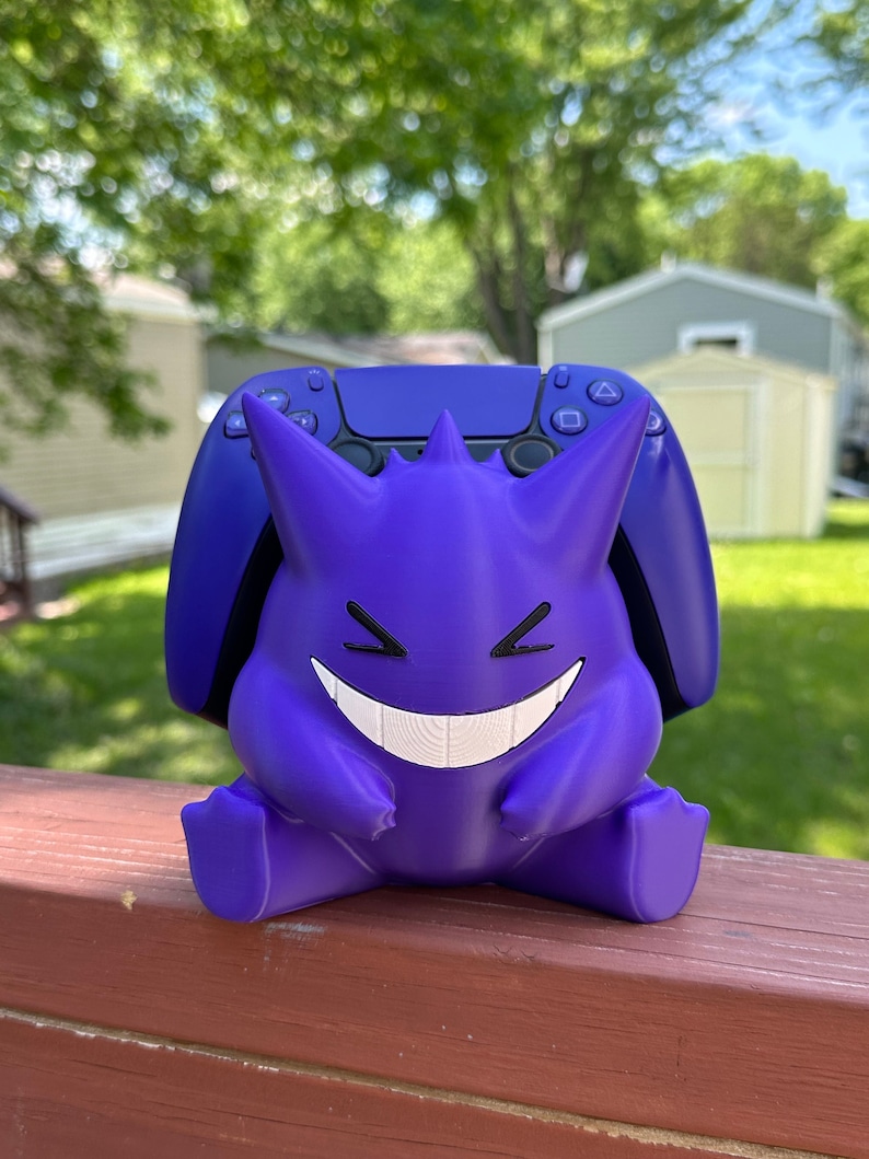 Peut inclure: Un support de manette violet en forme de Gengar avec une manette PlayStation 5 violette assortie. Le support a un visage souriant et est pos&eacute; sur une surface en bois. L'arri&egrave;re-plan est une sc&egrave;ne ext&eacute;rieure floue.