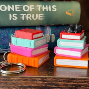 May include: A collection of colorful miniature book stacks, each with a different color spine and pages. A silver keychain is in the foreground. The text "NONE OF THIS IS TRUE" is visible on a book in the background.
