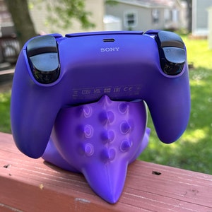 Peut inclure: Une manette de PlayStation 5 violette repose sur un support tentaculaire violet et &eacute;pineux assorti. La manette a des boutons noirs et le logo Sony. Le support est sur une surface en bois, avec un arri&egrave;re-plan ext&eacute;rieur flou.