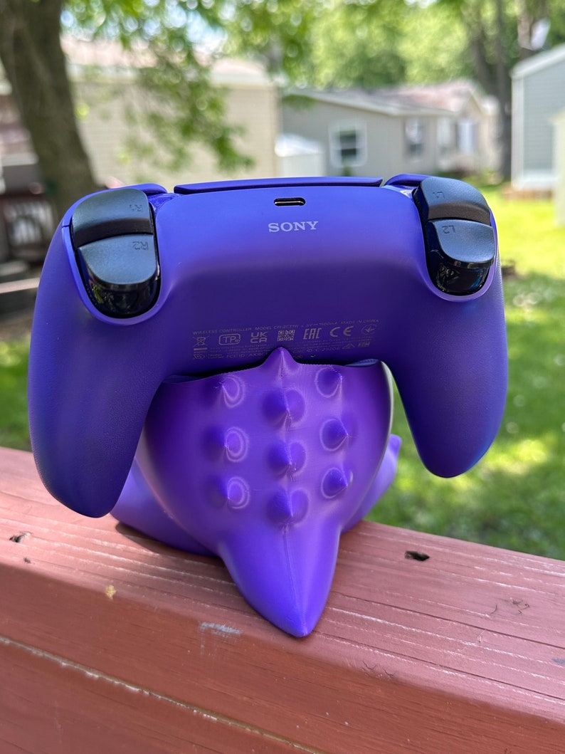 Peut inclure: Une manette de PlayStation 5 violette repose sur un support tentaculaire violet et &eacute;pineux assorti. La manette a des boutons noirs et le logo Sony. Le support est sur une surface en bois, avec un arri&egrave;re-plan ext&eacute;rieur flou.