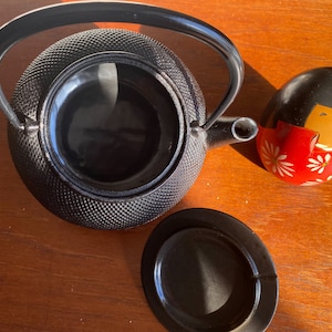 Puede incluir: Tetera de hierro fundido negro con una superficie texturizada y una tapa. La tetera tiene un asa y un pico. Una muñeca de madera negra y roja está en el fondo.